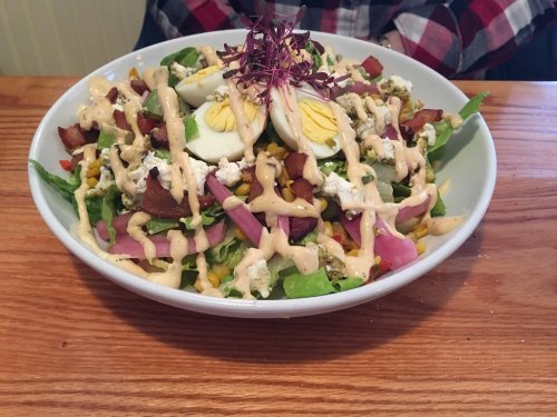 A colorful salad topped with sliced eggs, bacon, cheese, and drizzled with a creamy dressing, served in a bowl.