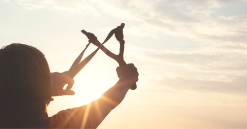 The silhouette of a young boy pulling at a slingshot toward the sky at sunset.