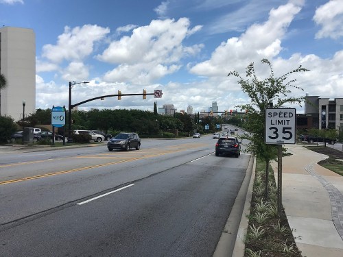 A wide road with a speed limit sign, cars driving, and a clear blue sky with clouds in the background.