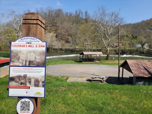 Sign for Cochran's Mill & Dam in Marion County, with a grassy area, picnic tables, and a river in the background.