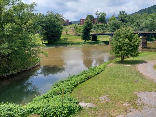 A serene river scene with green banks, trees, and a bridge in the background under a cloudy sky.