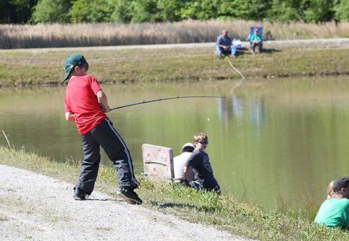 A child in a red shirt casts a fishing line by a pond, while others fish nearby on a sunny day.