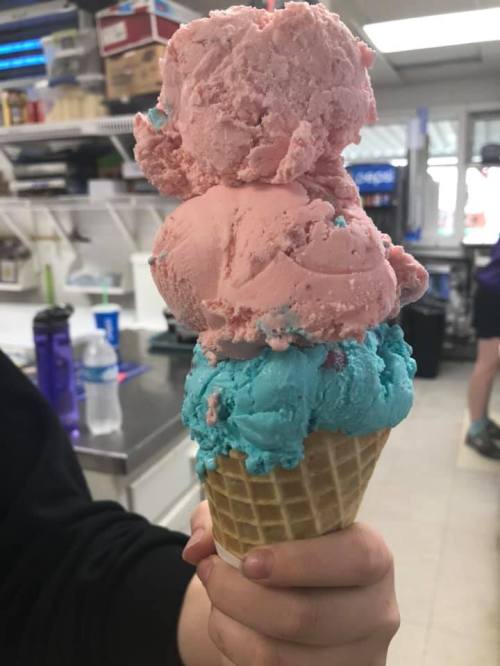 A hand holding a cone with three scoops of ice cream: pink, blue, and a mix of both on top.