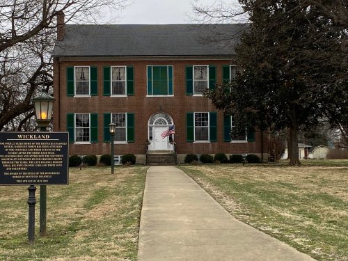 Historic brick building with green shutters, a flag, and a sign in front, surrounded by a grassy area.