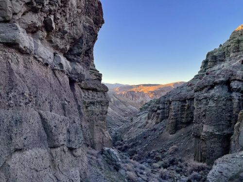 A narrow canyon with steep, rocky walls and a clear sky at dawn, showcasing rugged terrain and distant mountains.