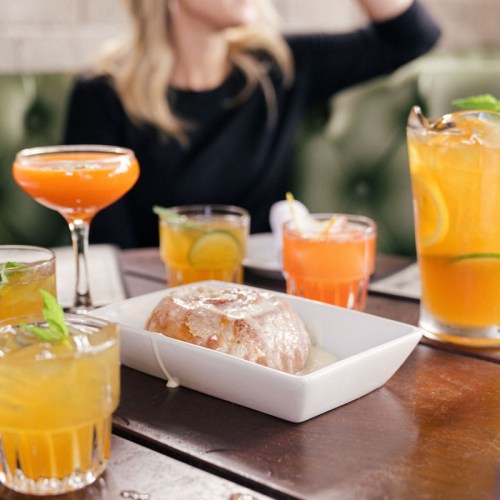 A table with colorful cocktails and a dish of food, with a blurred figure in the background.