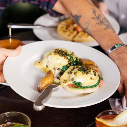 A person serves a plate of eggs Benedict with spinach and crispy potatoes, garnished with green herbs.