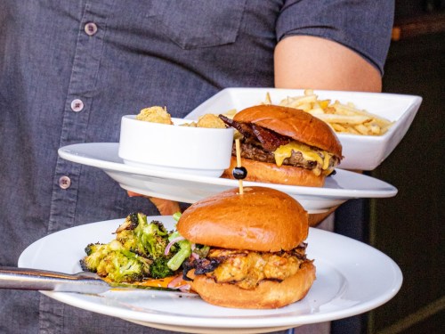 A person holds two plates: one with a burger and broccoli, the other with a burger and fries.