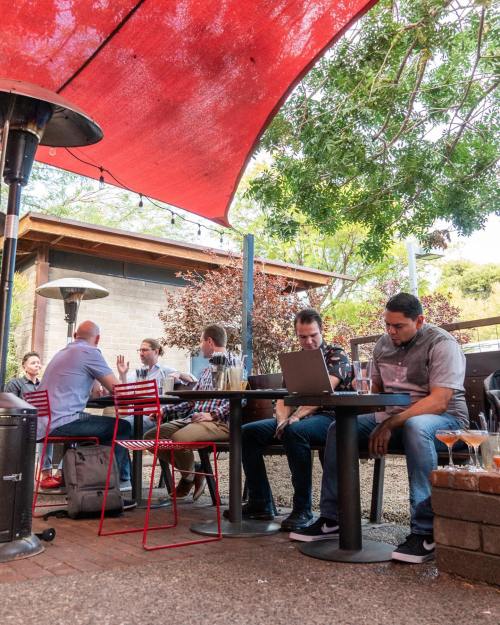 A group of people sitting at an outdoor café under a red canopy, engaged in conversation and working on laptops.
