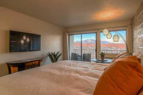 Cozy bedroom with a large bed, TV, and balcony view of mountains, featuring modern decor and warm lighting.