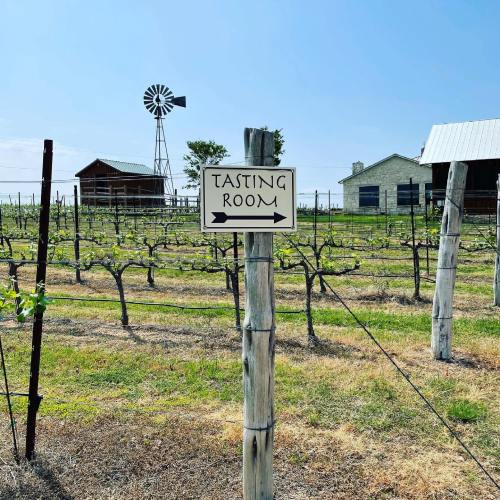 A sign pointing to a tasting room in a vineyard, with a windmill and buildings in the background.