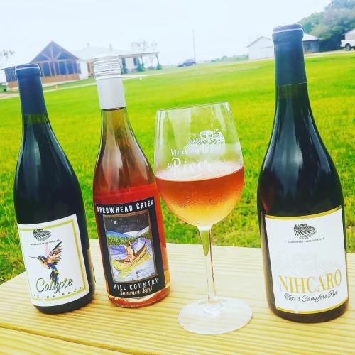 Three wine bottles and a glass of rosé on a wooden table, with a green field and building in the background.