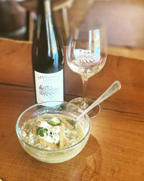 A bowl of soup with toppings, accompanied by a bottle of wine and a wine glass on a wooden table.