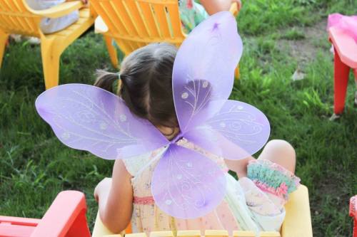 A child sits on a chair, wearing large purple butterfly wings, with her back to the camera, surrounded by colorful chairs.