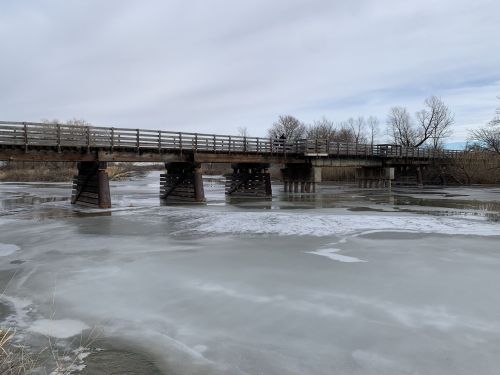 A wooden bridge spans a frozen river, surrounded by bare trees and a cloudy sky. Ice covers the water's surface.