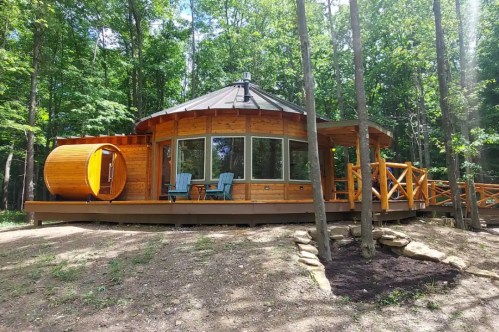 A round wooden cabin surrounded by trees, featuring a deck, two chairs, and a unique circular entrance.