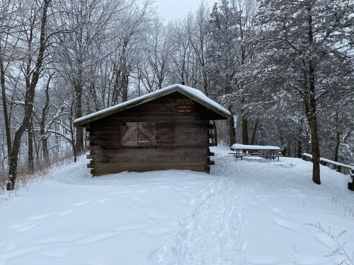 A snow-covered cabin surrounded by trees, with a picnic table nearby and a path leading through the snow.