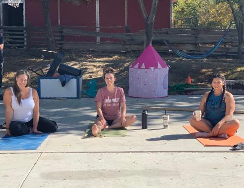 Three women practicing yoga outdoors on colorful mats, with a pink play tent and trees in the background.