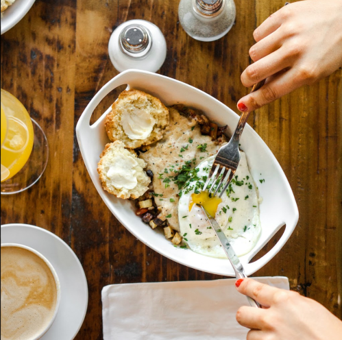 A hand holds a fork and knife over a dish with eggs, biscuits, and herbs, alongside a drink and coffee on a wooden table.