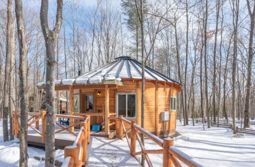A cozy wooden cabin with a circular design, surrounded by snow-covered trees and a clear blue sky.