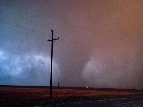 A dark, stormy sky looms over a flat landscape with power lines, hinting at a tornado in the distance.
