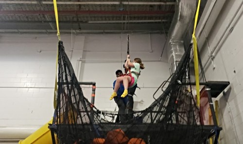 A child and an adult are on a zip line above a safety net in an indoor play area.