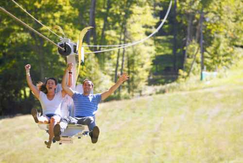 A man and woman joyfully ride a chairlift, raising their arms in excitement against a backdrop of green trees.