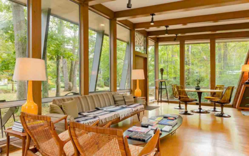 A modern living room with large windows, wooden beams, a sofa, and a dining table surrounded by greenery outside.