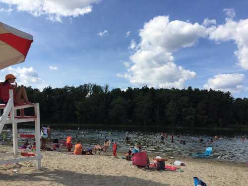 A sandy beach scene with people swimming and relaxing by a lake, under a blue sky with fluffy clouds. A lifeguard watches from a chair.