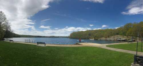 A serene lake scene with benches, green grass, and trees under a partly cloudy sky.