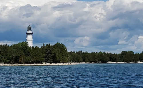 A lighthouse stands on a shoreline surrounded by trees, with a blue sky and clouds above. Water in the foreground.