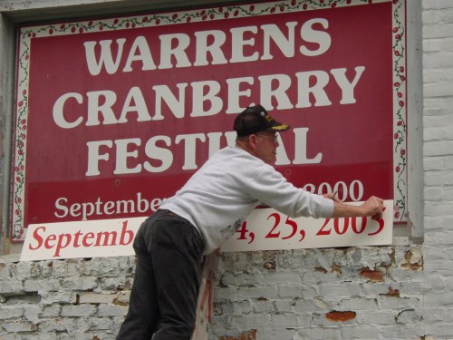 A man hangs a sign for the Warrens Cranberry Festival on a brick wall, adjusting the date to September 23, 2003.