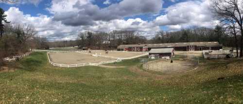 A panoramic view of a horse farm with stables, riding arenas, and a cloudy sky in the background.