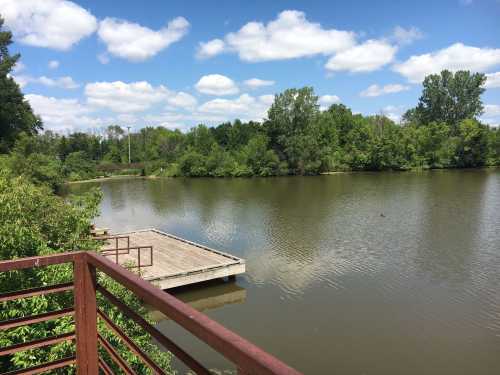 A serene view of a calm lake surrounded by lush greenery and a wooden dock under a partly cloudy sky.