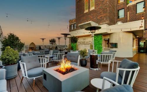 Rooftop lounge with modern seating, a fire pit, and greenery, overlooking a sunset skyline.