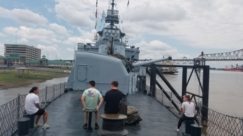 Three people sit on a ship's deck, facing a large naval gun, with a city skyline and cloudy sky in the background.