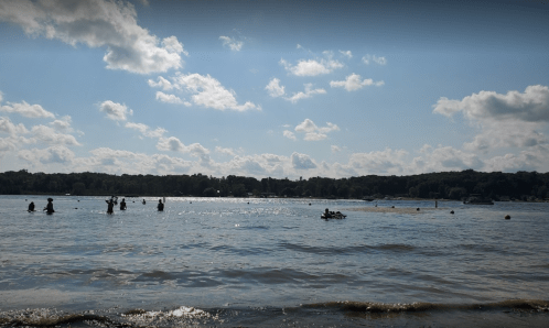People enjoying a sunny day at the lake, with some swimming and others on jet skis, under a partly cloudy sky.