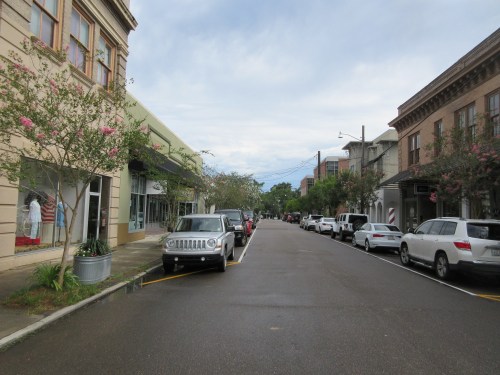 A quiet street lined with parked cars and shops, featuring trees and a cloudy sky overhead.
