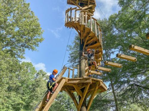 Two children navigate a wooden climbing structure in a forested area, with a spiral staircase and suspended platforms.