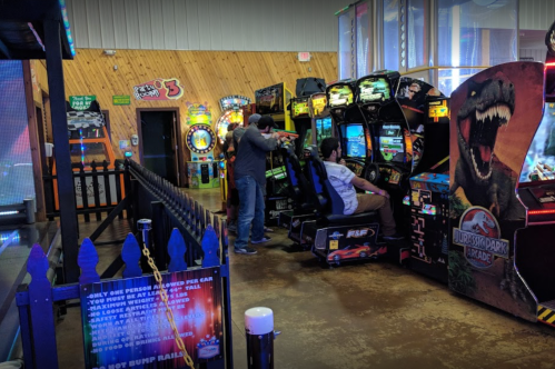 Two people play arcade games in a colorful game room filled with various machines and a Jurassic Park-themed cabinet.