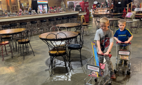 Two young boys push toy shopping carts in a cozy, rustic café with empty tables and festive decorations.
