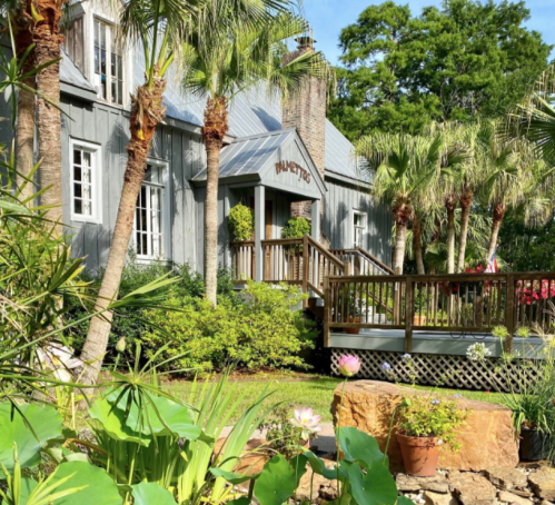 A charming gray house with a wooden deck, surrounded by palm trees and lush greenery, featuring colorful flowers in the foreground.