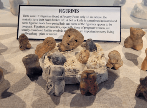 A display of ancient figurines from Poverty Point, some with broken heads, on a stone base with an informational sign.