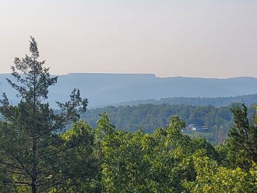 A hazy landscape featuring distant mountains, lush green trees in the foreground, and a clear blue sky.