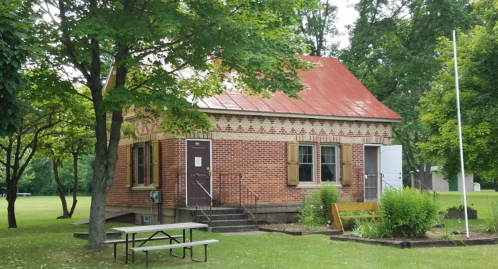 A small brick building with a red roof, surrounded by trees and a grassy area, featuring picnic tables and benches.