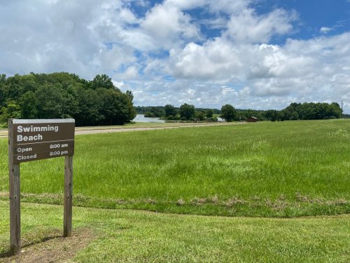 A grassy area with a sign for a swimming beach, under a partly cloudy sky, near a lake in the background.
