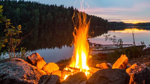 A campfire burns brightly by a tranquil lake at sunset, surrounded by trees and rocks.
