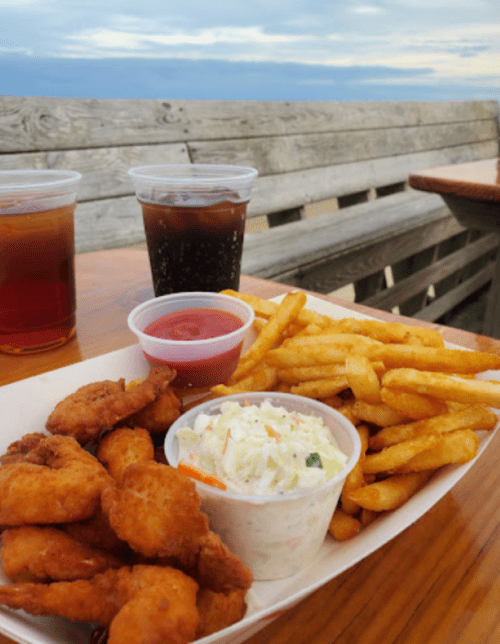 A plate of fried shrimp, coleslaw, and fries, with two cups of iced drinks on a wooden table by the beach.