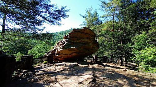A large rock formation sits on a rocky ledge, surrounded by trees and a wooden railing under a clear blue sky.