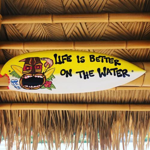 A colorful surfboard with a tiki design and the phrase "Life is better on the water" displayed on a bamboo backdrop.